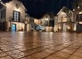 Night view of white stucco buildings with string lights, a wet cobbled plaza, and warmly lit doors and windows in a village square.