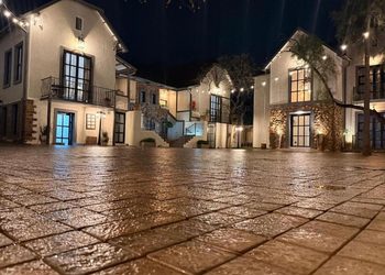 Night view of white stucco buildings with string lights, a wet cobbled plaza, and warmly lit doors and windows in a village square.