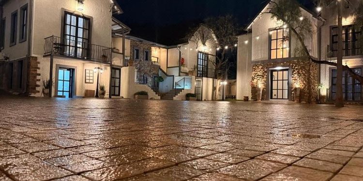 Night view of white stucco buildings with string lights, a wet cobbled plaza, and warmly lit doors and windows in a village square.